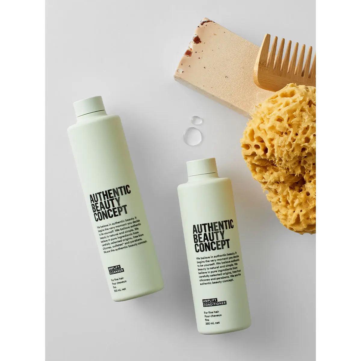 Two bottles of Authentic Beauty Concept Amplify Cleanser with a wooden comb and natural sponge on a light background.