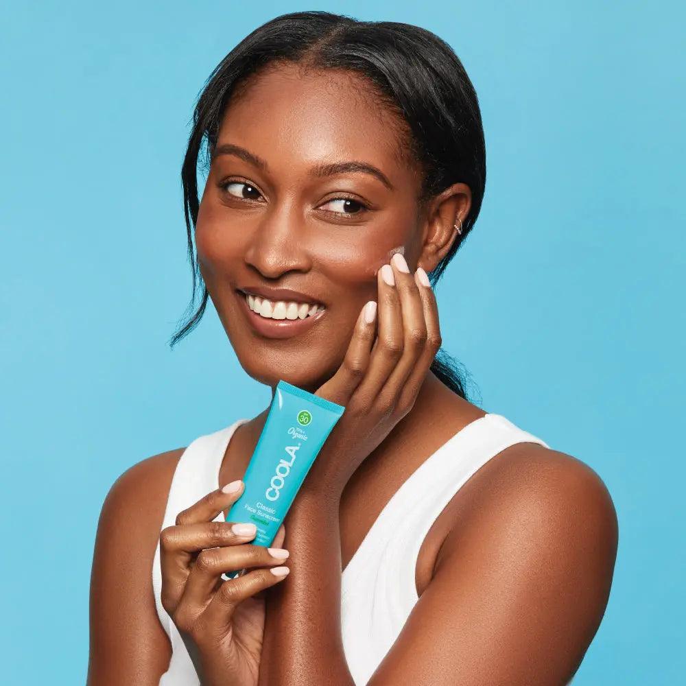 Smiling woman holding Classic Face Organic Sunscreen Lotion SPF 30 with a blue background, showcasing radiant skin protection.