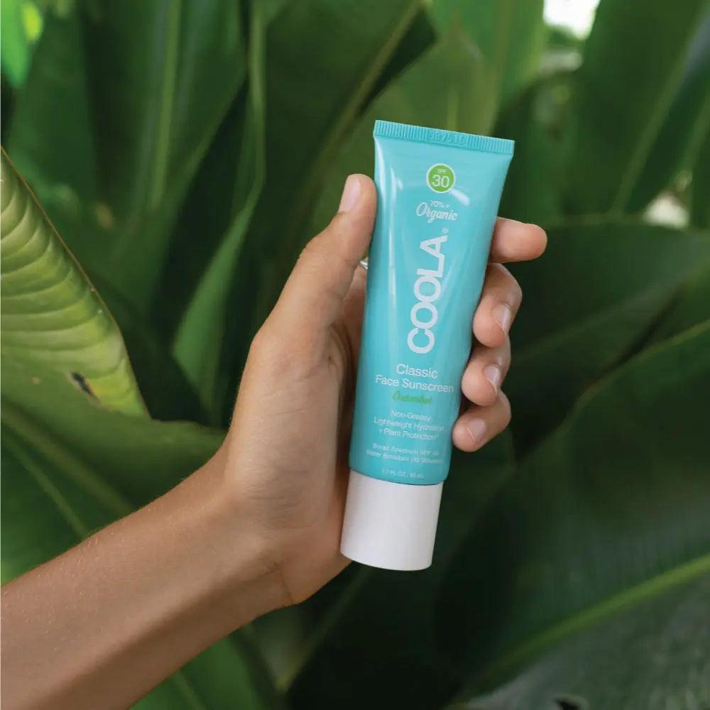 Hand holding COOLA Classic Face Sunscreen Lotion SPF 30 in front of green foliage, promoting organic skin protection.