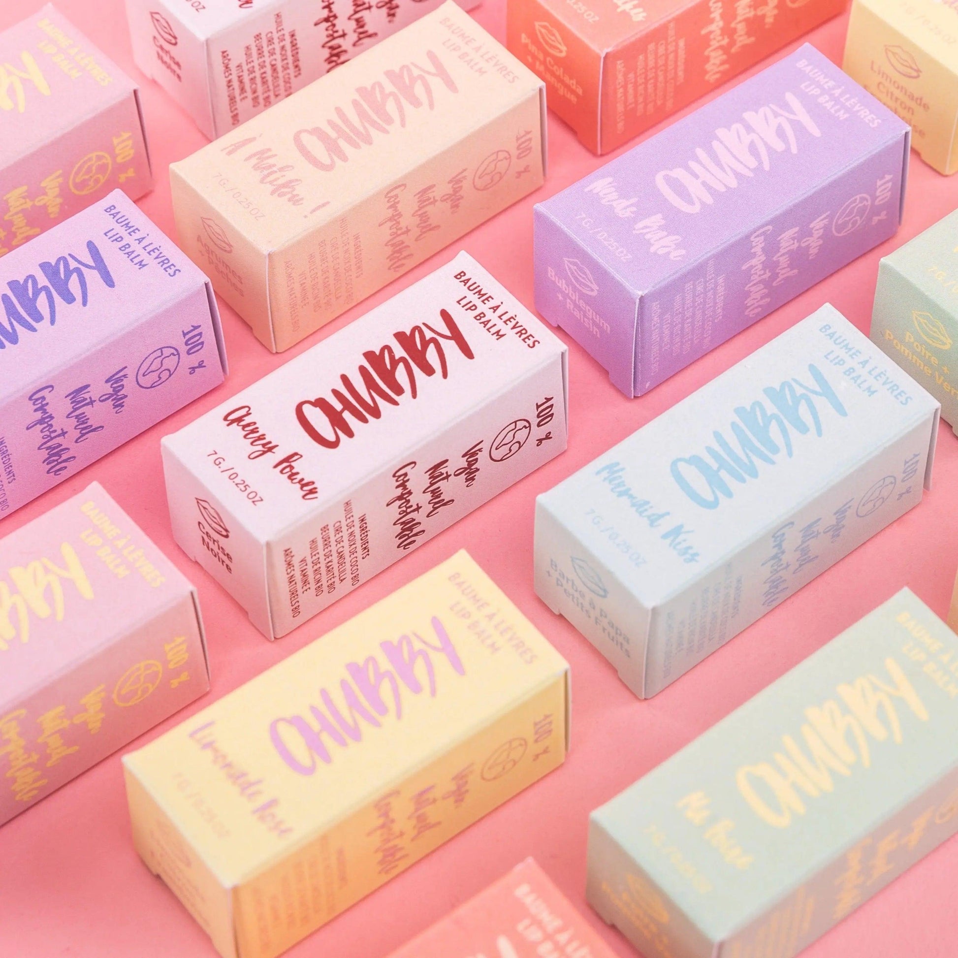 Colorful packaging of compostable vegan lip balms displayed on a pink background, showcasing various flavors and eco-friendly branding.