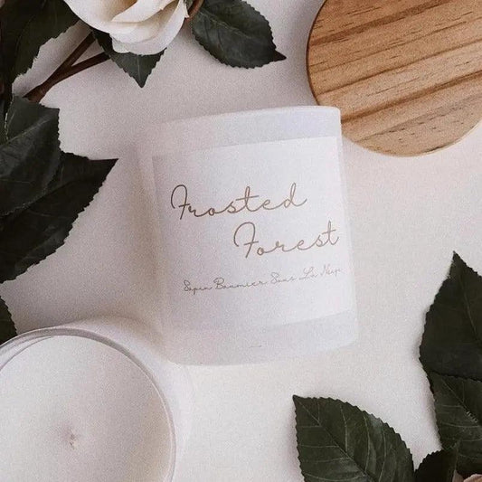 Frosted Forest candle with elegant label on a white jar, surrounded by greenery and wooden lid, showcasing natural aesthetics.