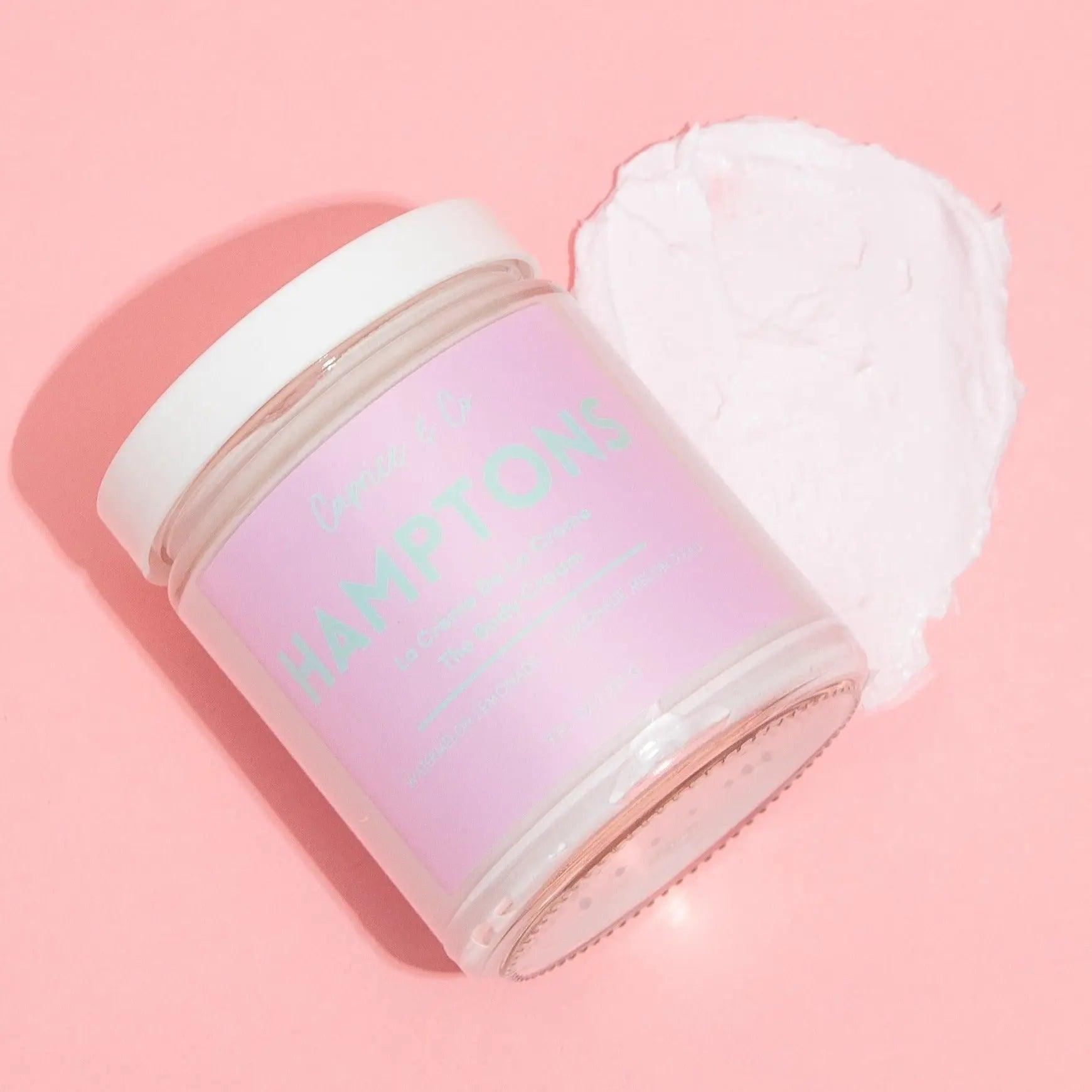 Hamptons body cream jar on pink background with cream texture beside it, showcasing its moisturizing properties.