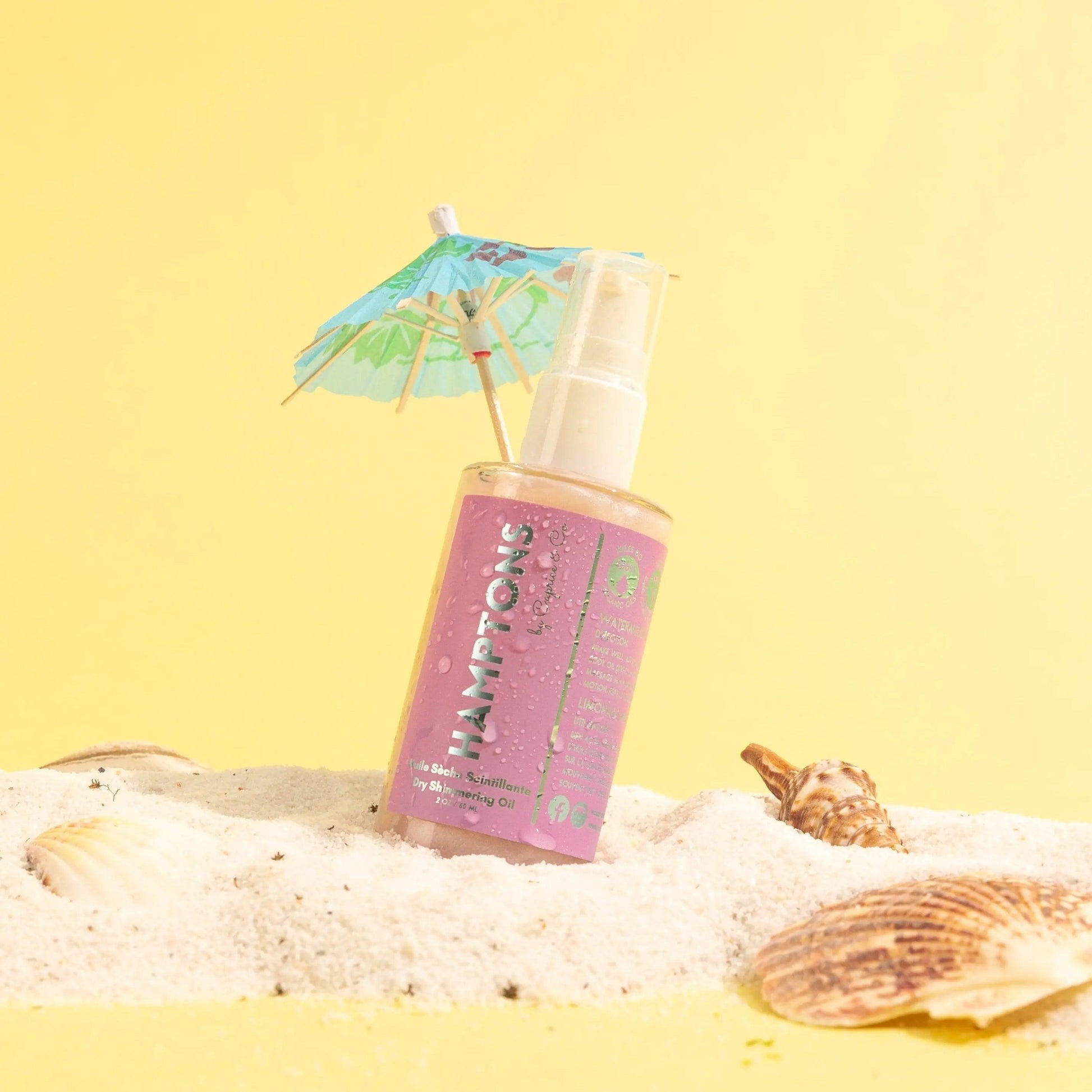Hamptons Dry Shimmering Oil with pink packaging on sandy beach with seashells and a mini umbrella, vibrant yellow background.