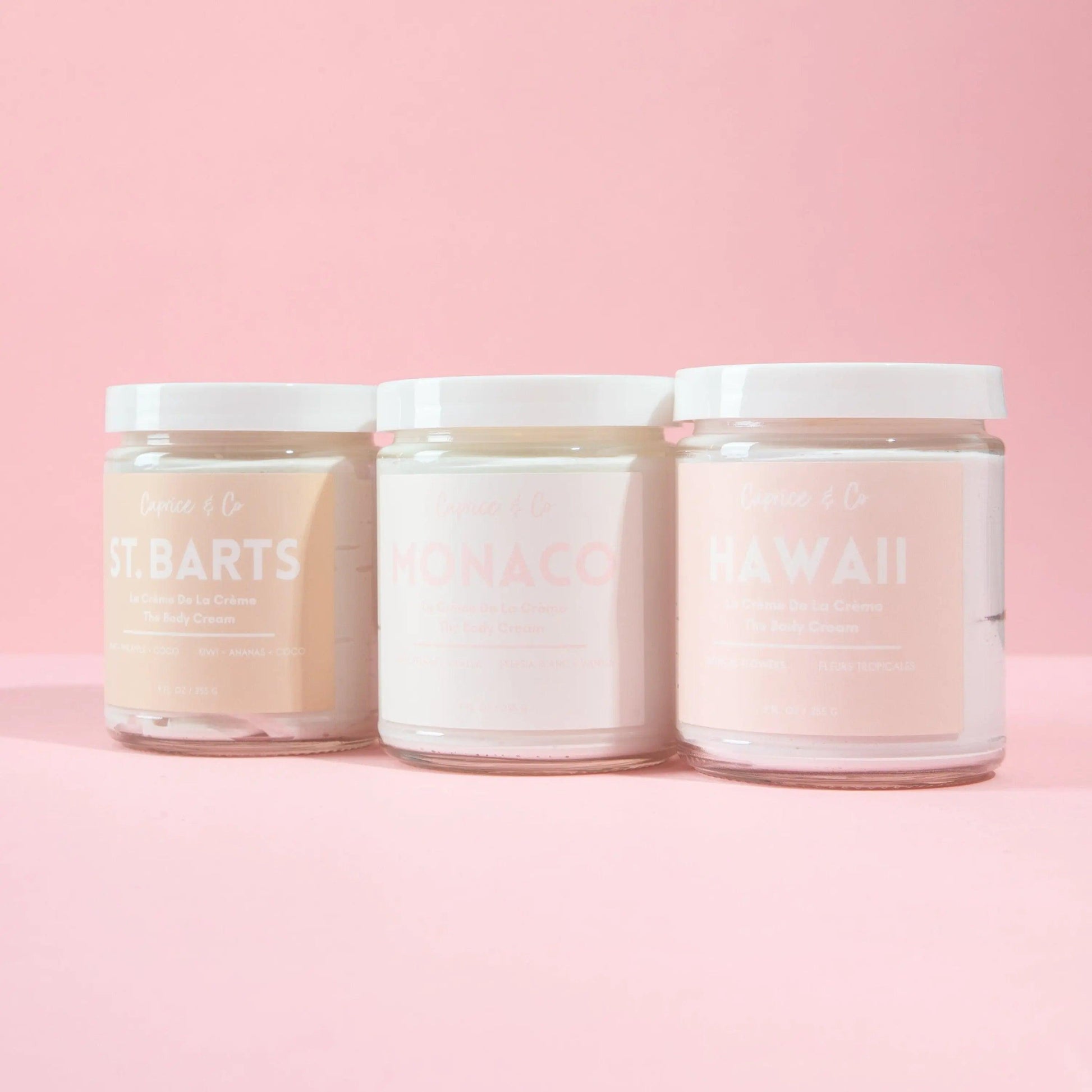 Trio of body creams in glass jars: St. Barts, Monaco, and Hawaii on a pink background. Hydrating and luxurious scents.