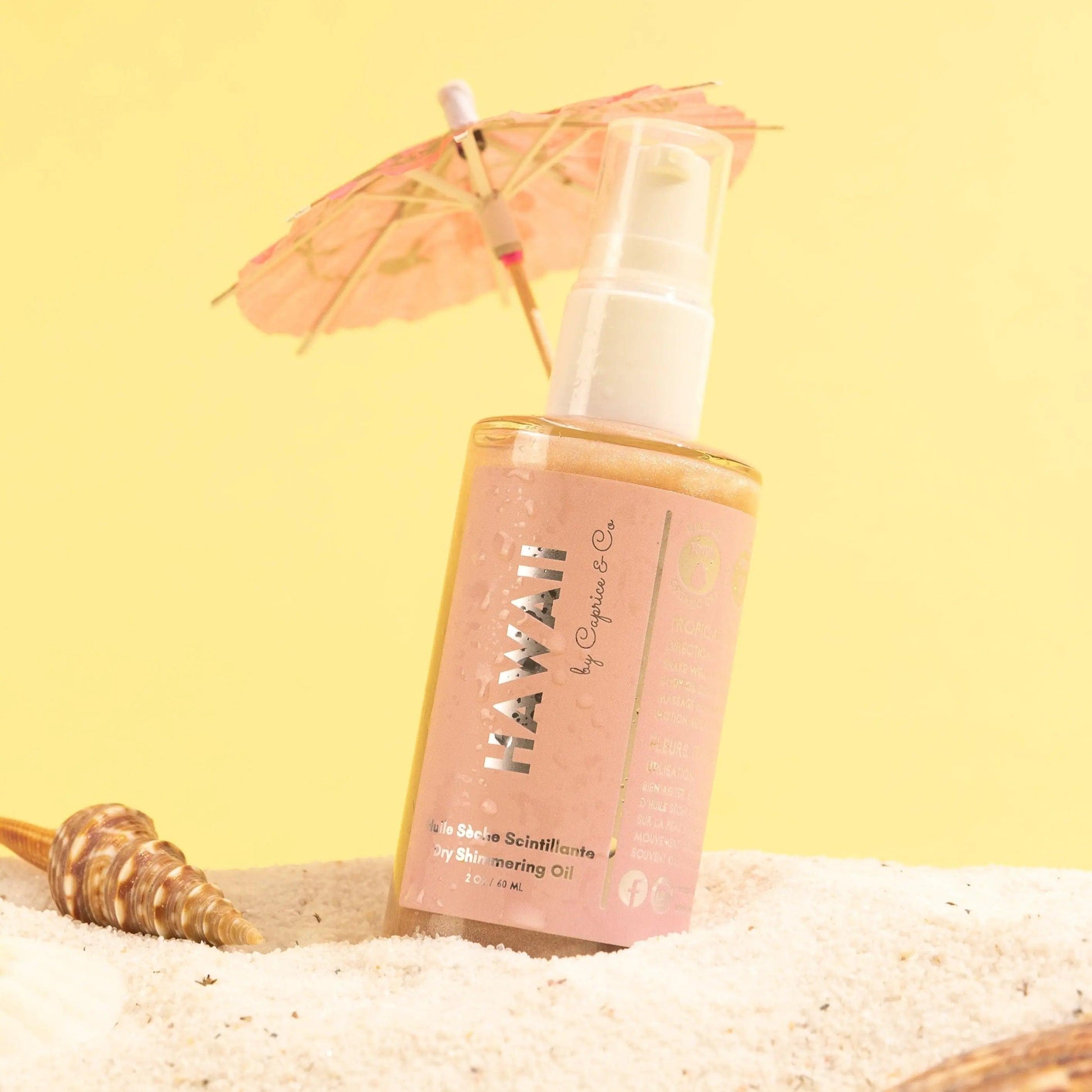 Hawaii Dry Shimmering Oil bottle on sandy beach with tropical umbrella, showcasing rose gold shimmer and organic ingredients.