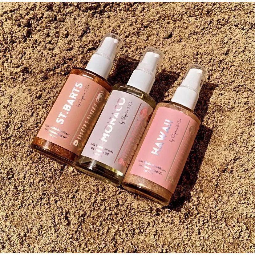 Three bottles of dry shimmering oil labeled St. Barts, Monaco, and Hawaii, placed on sandy beach surface.