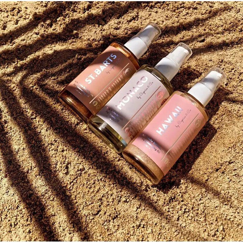 Three bottles of body oil, including Monaco, St. Bart's, and Hawaii, on sandy beach with shadows.