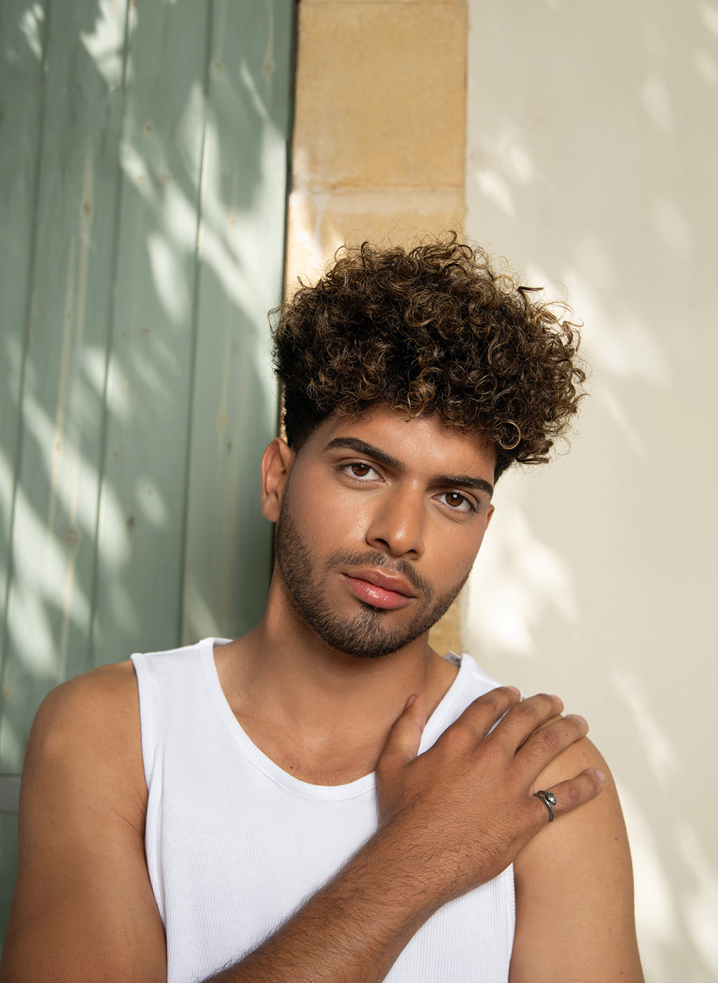 Young man with curly hair showcasing a stylish look against a rustic background, embodying confidence and modern fashion.