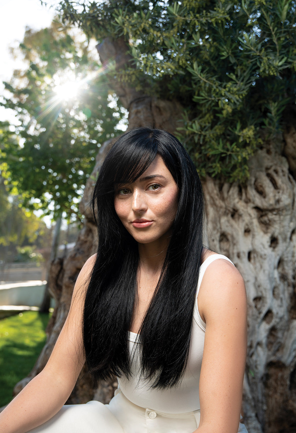 Woman with long, healthy black hair sitting by a tree, showcasing radiant hair in natural sunlight.
