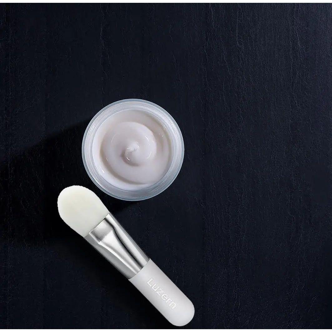 Luxurious skincare cream in a glass jar with a white applicator brush on a black background