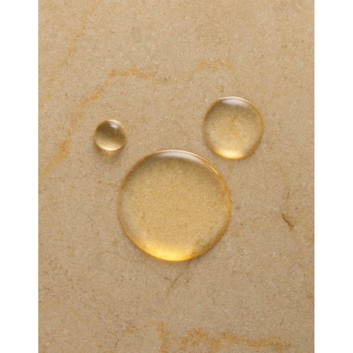 Close-up of three glistening drops of argan oil on a beige surface, highlighting its rich texture and hydration benefits.