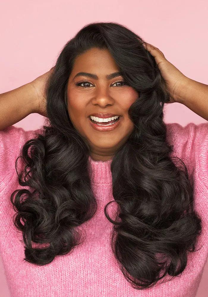 Smiling woman with long, voluminous black hair, wearing a pink sweater, showcasing healthy, shiny curls.