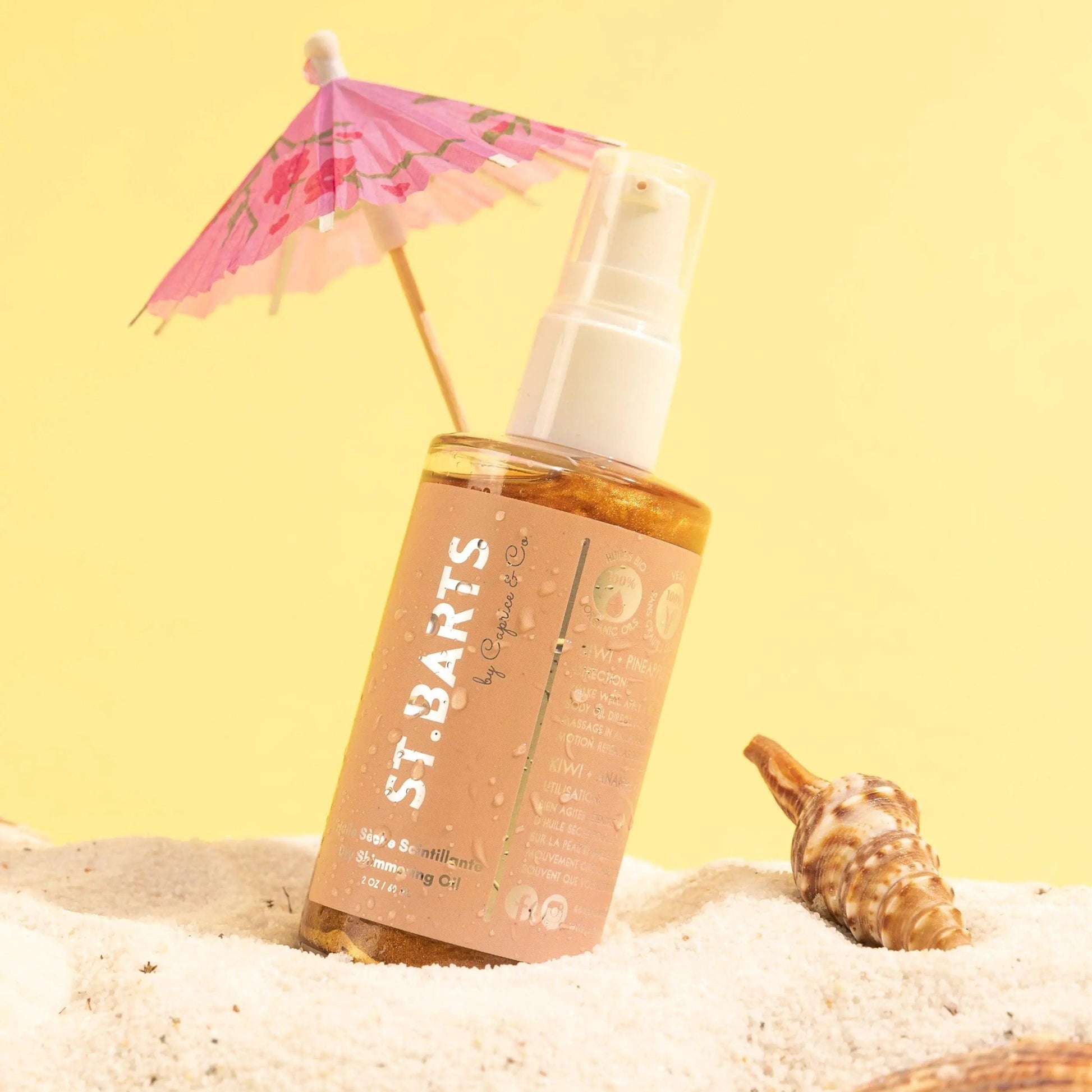 St. Barts Dry Shimmering Oil bottle on sandy beach with a pink umbrella, showcasing a bronze color and tropical vibe.