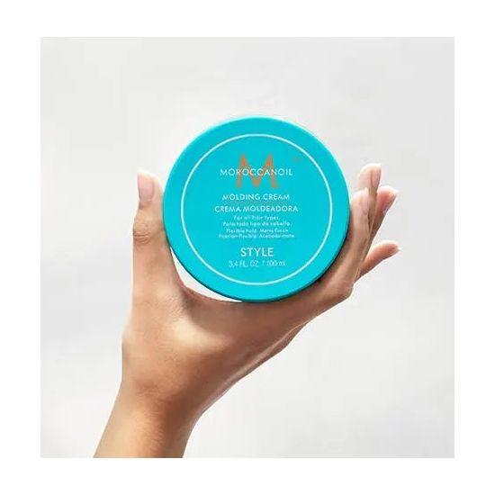 Hand holding a jar of Molding Cream from Moroccanoil, showcasing its vibrant blue packaging and rich texture.