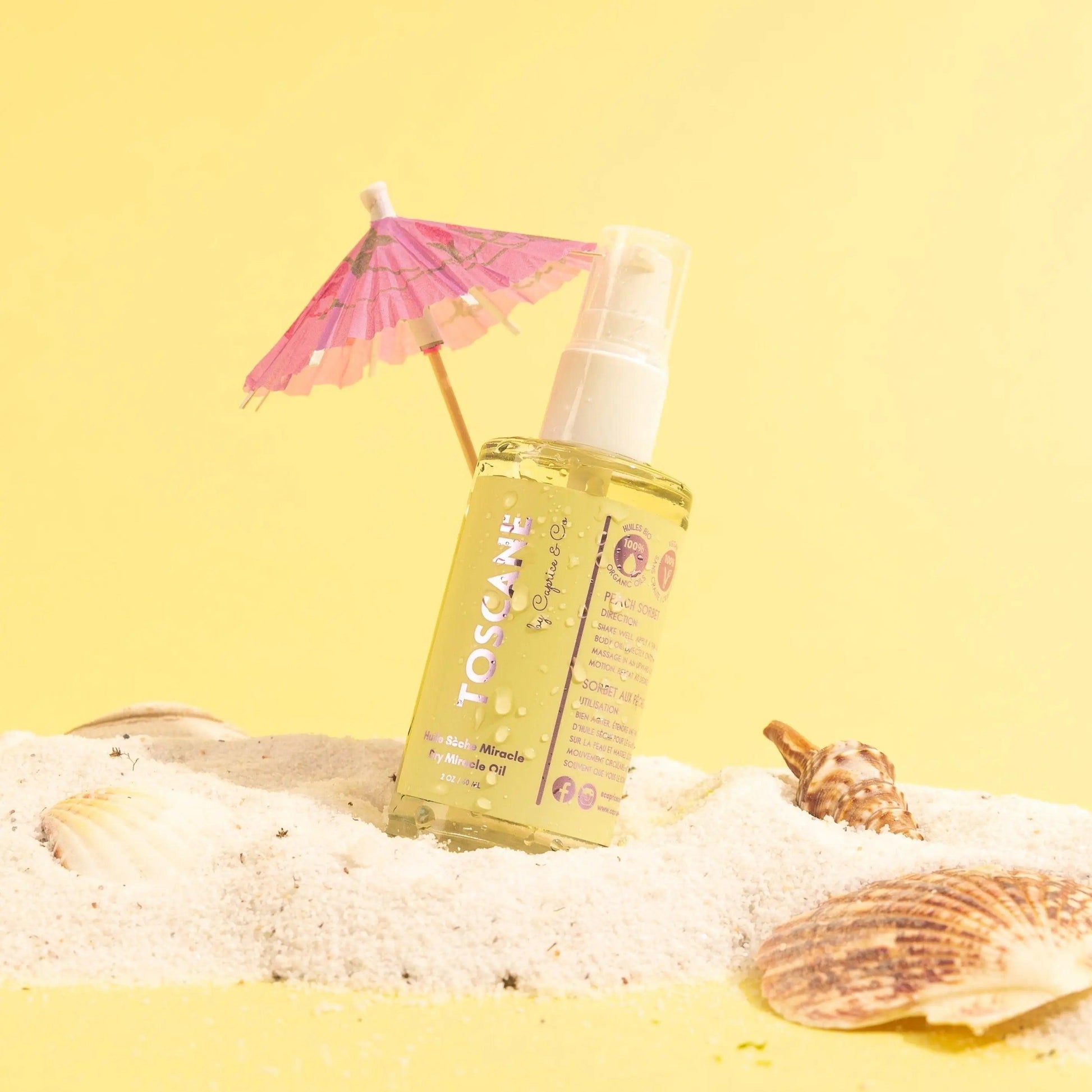 Toscane Dry Miracle Oil in a beach setting, featuring peach sorbet scent with organic ingredients, surrounded by seashells and sand.