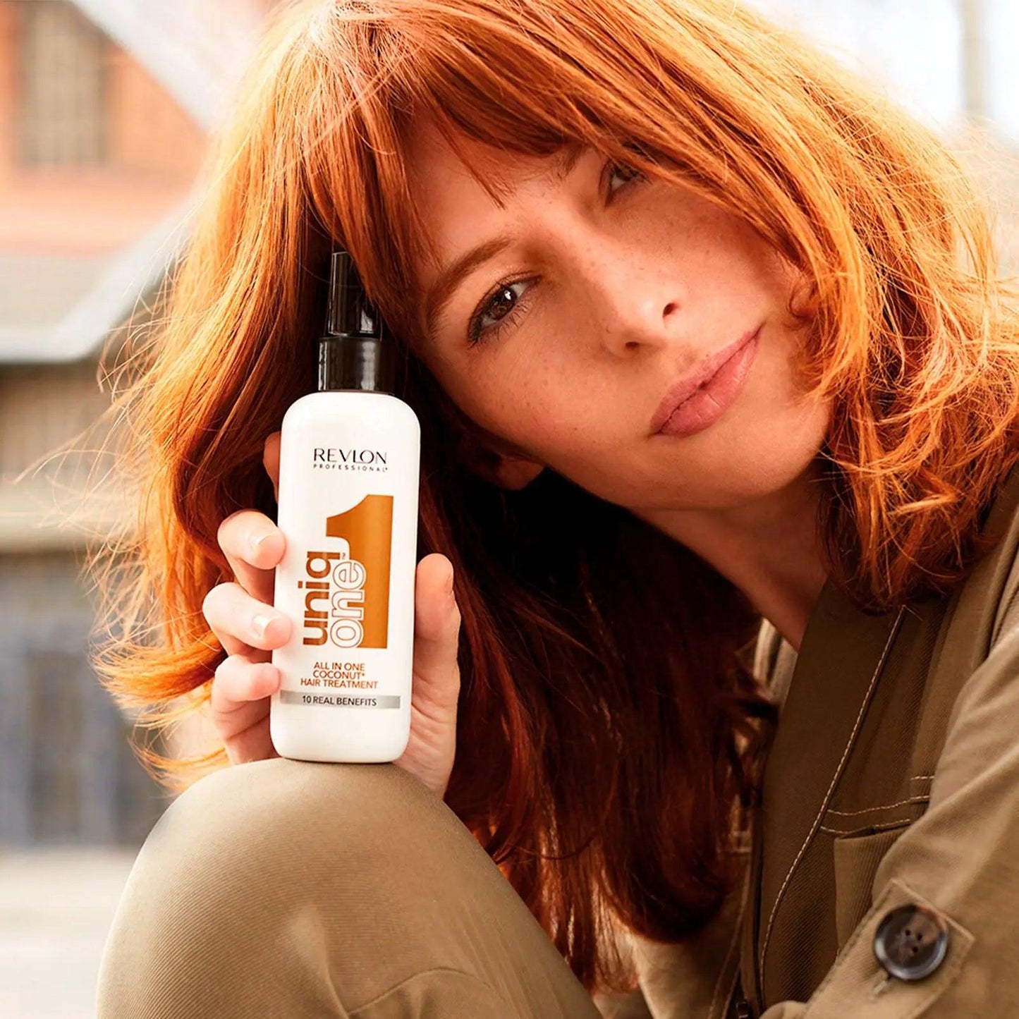 Woman with red hair holding Revlon Uniq One hair treatment bottle outdoors.