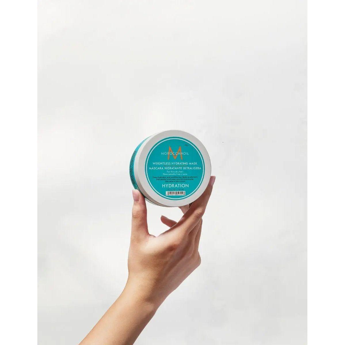 Person holding Moroccanoil Weightless Hydrating Mask for dry hair, featuring its turquoise packaging and hydration benefits.