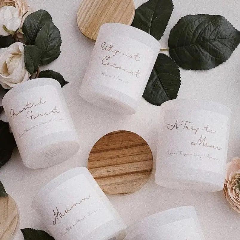Collection of natural candles including "Why Not Coconut," with wooden lids and surrounded by greenery and flowers.