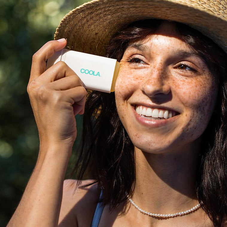 Woman applying Classic Organic Sunscreen Stick SPF 30 Tropical Coconut outdoors, smiling in sunlight with a straw hat.