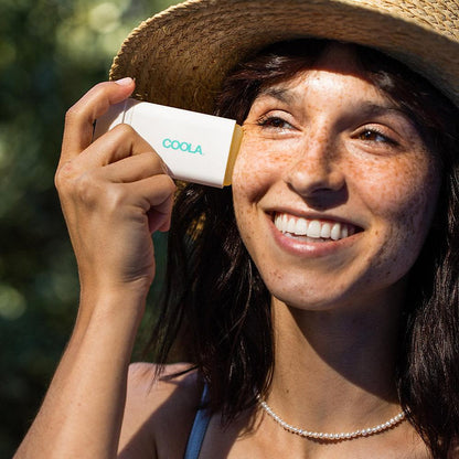 Woman applying Classic Organic Sunscreen Stick SPF 30 Tropical Coconut outdoors, smiling in sunlight with a straw hat.