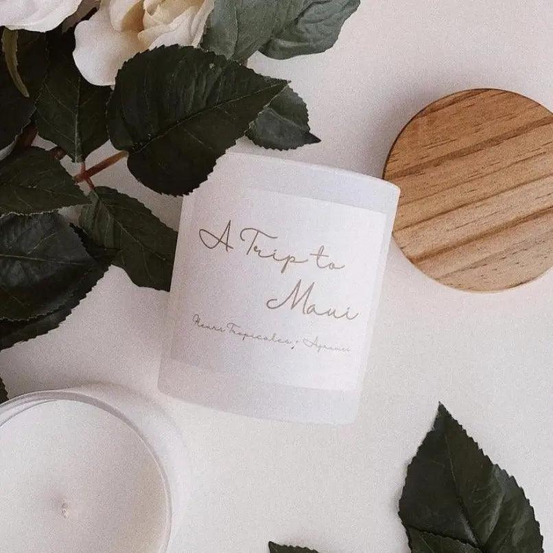 A Trip to Maui candle with tropical flowers, surrounded by leaves and a wooden lid on a neutral background.