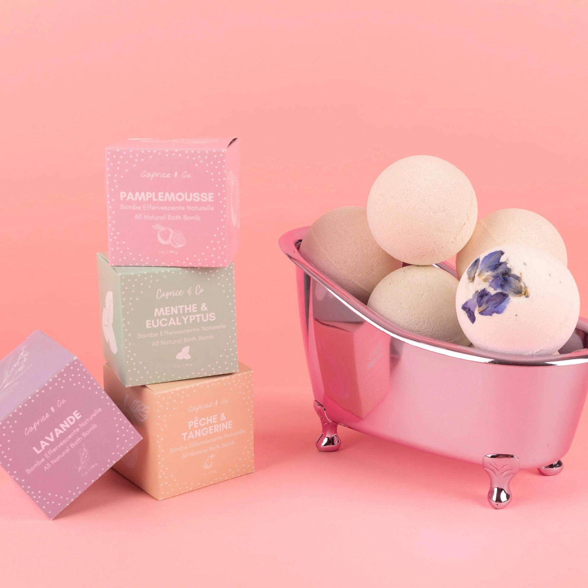 All natural bath bombs in a silver tub with colorful packaging on a pink background.