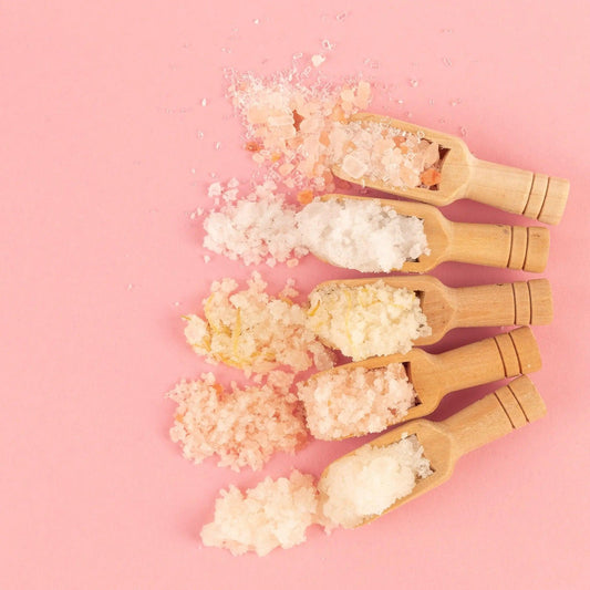 Assorted natural bath salts in wooden scoops on a pink background, showcasing pink Himalayan and soothing Epsom salts.