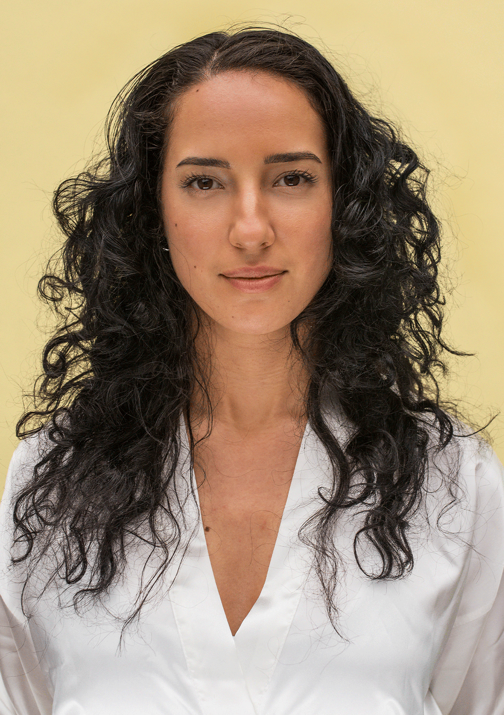 Woman with defined, bouncy curls wearing a white blouse against a yellow background, showcasing vibrant curly hair.
