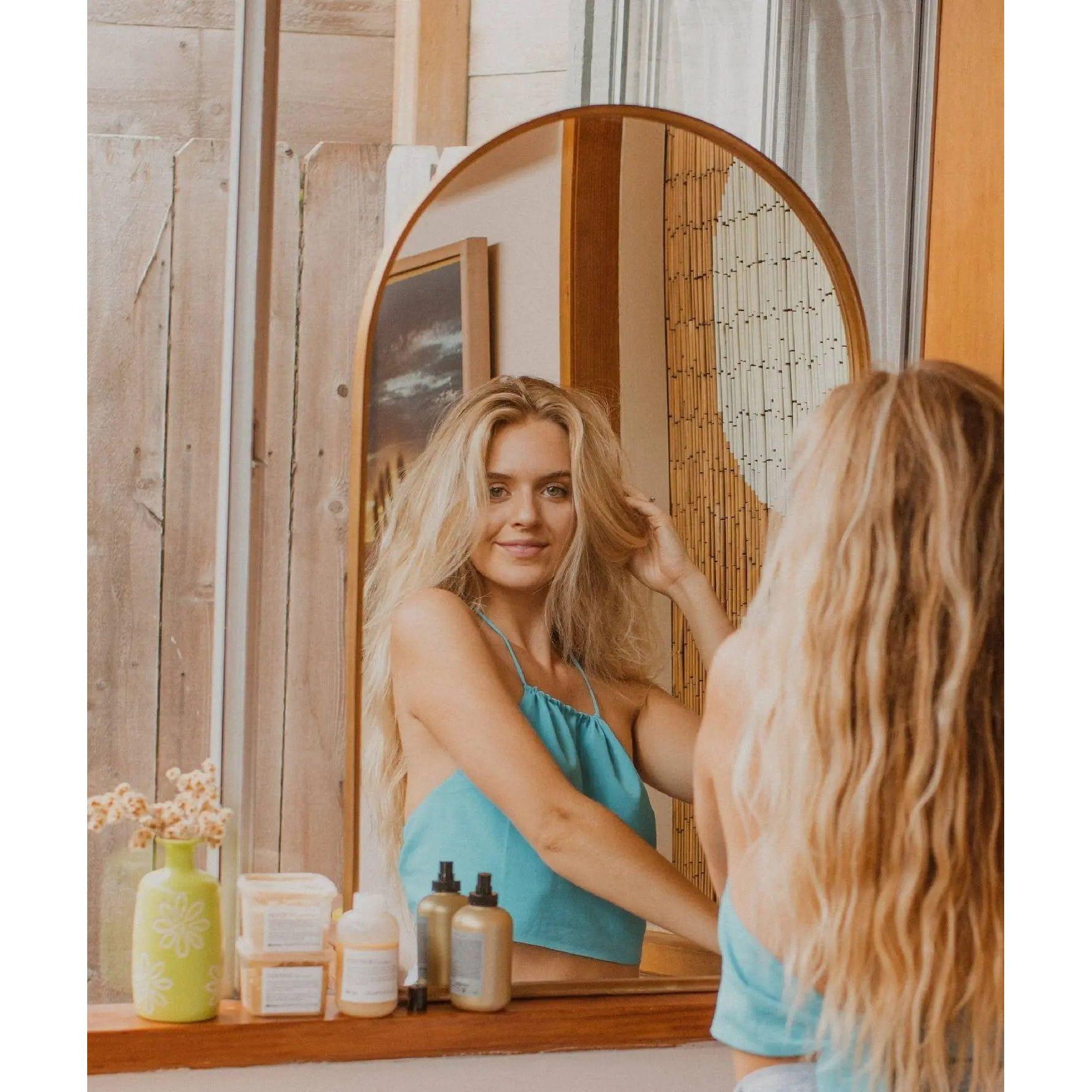 Woman with long, wavy hair admiring her reflection, showcasing hair care products on the countertop in a cozy setting.