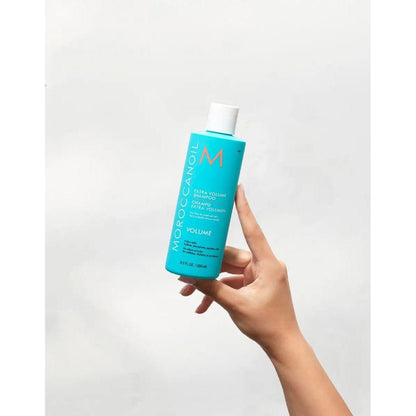 Hand holding Moroccanoil Extra Volume Shampoo bottle against a neutral background.