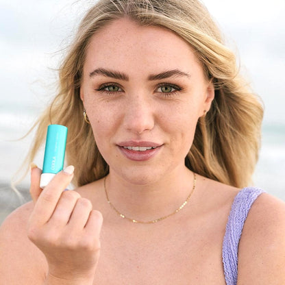 Woman holding Classic Liplux® Organic Lip Balm SPF 30, showcasing radiant skin and beachy backdrop.