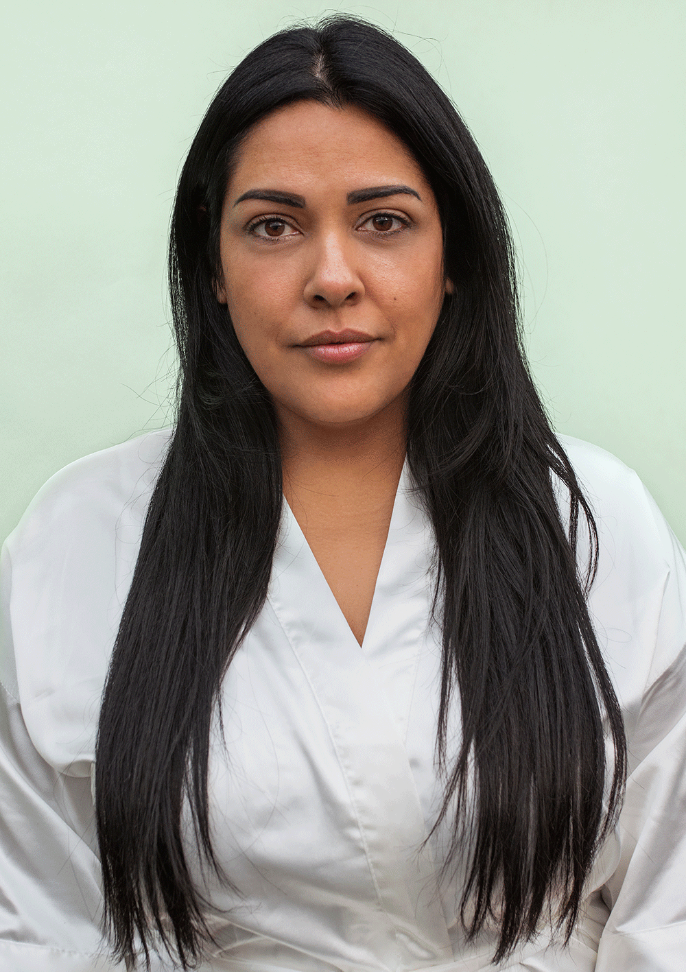 Woman with long, straight dark hair wearing a white robe against a light green background.