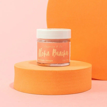 Aloha Beaches Lip Scrub jar on an orange pedestal, featuring Pina Colada and Mango flavors in a vibrant, stylish setting.