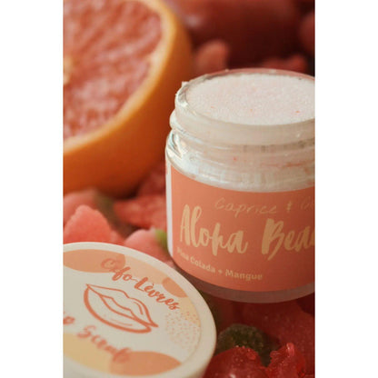 Aloha Beaches Lip Scrub in a jar surrounded by fruits, featuring Pina Colada and Mango flavors for gentle lip exfoliation.