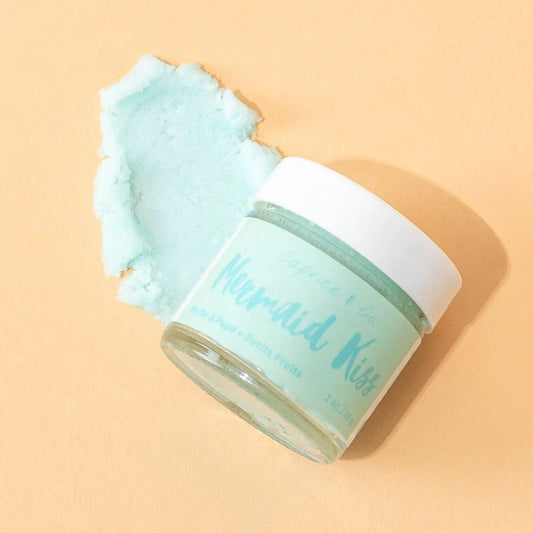 Mermaid Kiss lip scrub jar with cotton candy and berries flavor beside a pile of mint green scrub on a peach background.
