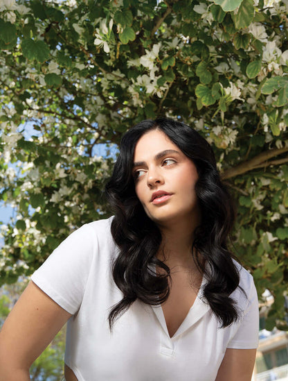 Woman with long, wavy black hair wearing a white shirt, posing in front of a blooming tree.