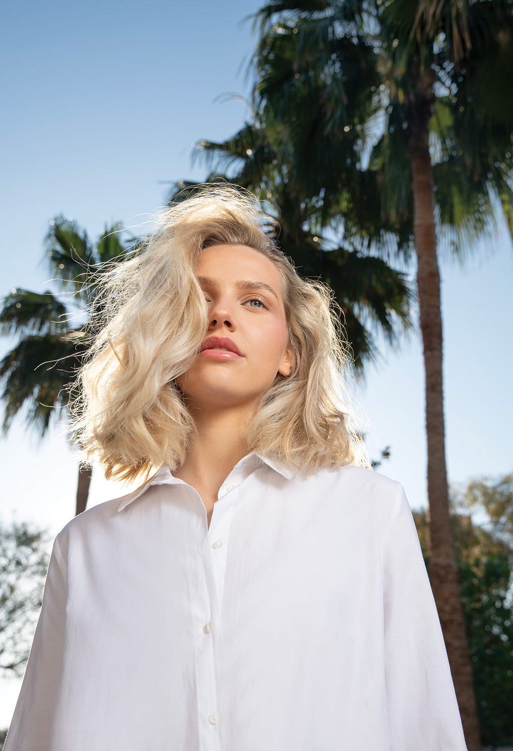 Woman with wavy blonde hair wearing a white shirt, standing among palm trees under a clear blue sky.