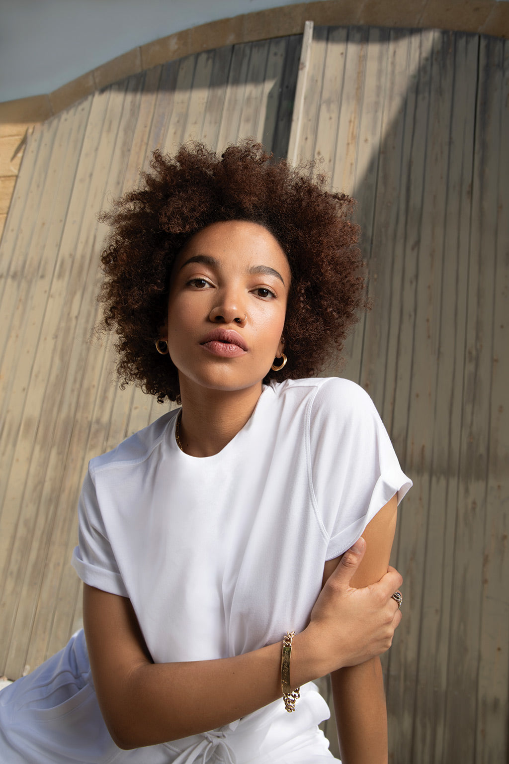 Model with curly hair wearing a white shirt, posing against a wooden background in natural light.