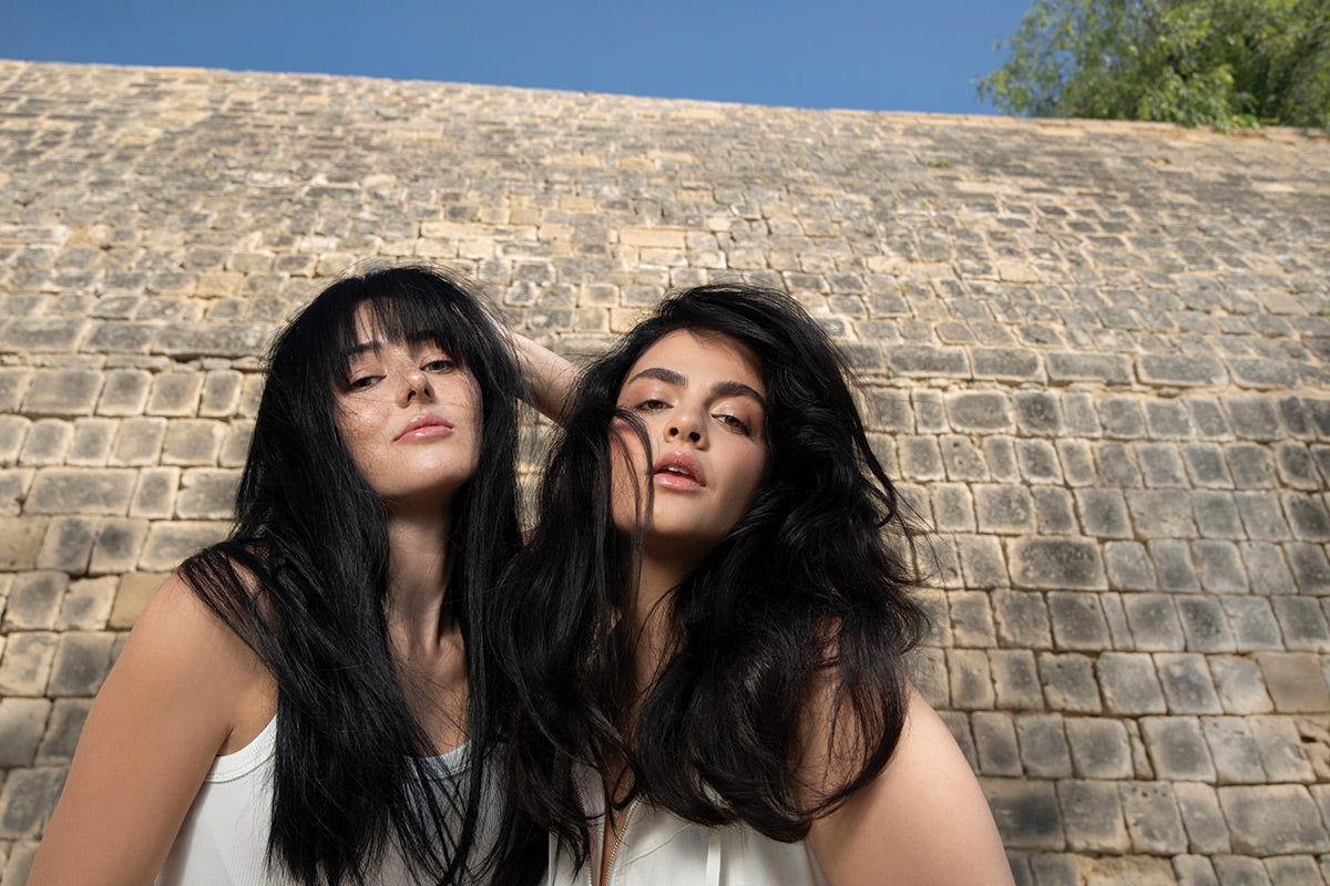 Two women showcasing voluminous, shiny hair styled with Neu Styling Mousse against a textured stone background.