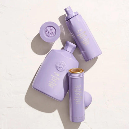 Oribe purple hair care products including a dry shampoo, treatment spray, and styling cream on a light background.