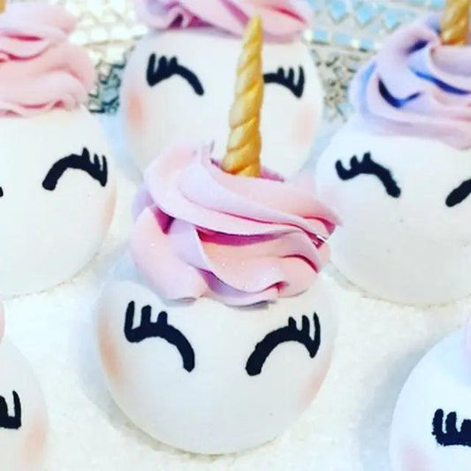 Adorable unicorn bath bombs with swirled pink and purple frosting, topped with golden horns, arranged on a shiny surface.
