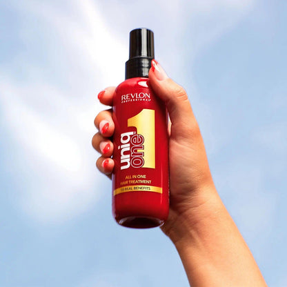 Hand holding Revlon Uniq One all-in-one hair treatment bottle against a blue sky.